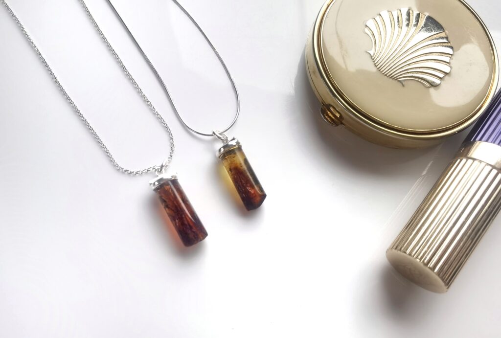 Jak dbać o biżuterię - 5 złych nawyków - Srebrne naszyjniki z podłużnymi wisiorami z ciemnego bursztynu bałtyckiego obok puderniczki w stylu vintage.