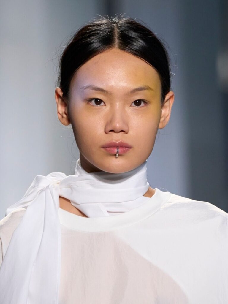 biżuteryjne trendy - Srebrny kolczyk w wardze typu kółko na twarzy modelki w białej stylizacji z ozdobnym wiązaniem pod szyją.